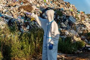An environmental worker dressed in protective gear surveys a landfill filled with waste while pointing at the surroundings under clear skies photo