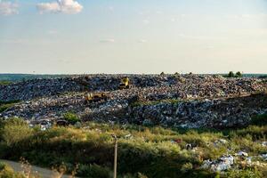 The landscape shows a sprawling landfill with heavy machinery working among heaps of refuse. Green vegetation partially surrounds the area, illuminated by afternoon sunlight photo