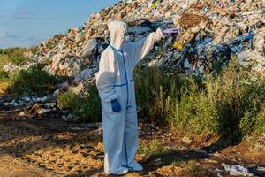 Wearing full protective clothing and gloves, a worker examines a large landfill, meticulously checking for any hazardous materials under the clear blue sky photo