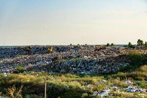 Bulldozers maneuver through a substantial landfill, compacting trash under a clear afternoon sky, surrounded by sparse vegetation and distant trees photo