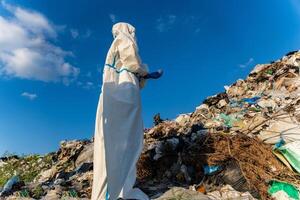 A waste management worker in protective clothing inspects a landfill filled with various types of debris under a sunny blue sky in the daytime photo