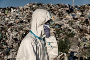 A person dressed in protective clothing and gloves examines a vast area filled with waste. The worker stands amidst piles of garbage, highlighting environmental concerns photo