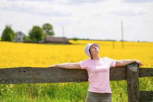 woman wearing a bucket hat leaning on an old wooden fence enjoying a warm summer day against a rustic farmstead surrounded by blooming rapeseed photo