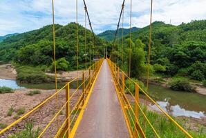 Yellow Traditional Iron Pedestrian Bridge Across Mountain River Against Mountain Backdrop In Vietnam photo