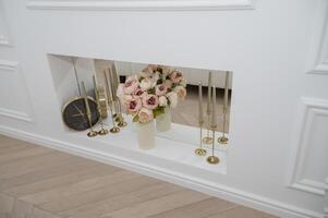 Interior details black clock, flowers, candles. white wall of the room with a built-in shelf. Clock, vase with pink flowers and golden candlesticks on a white shelf with a mirror surface photo
