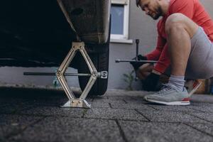 Close-up of man changing a car tire in front of his house. The vehicle is lifted with a jack, tools lie nearby. The scene captures a hands-on repair moment in daylight photo