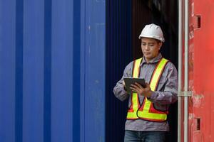 Asian Male Foreman Using Digital Tablet at Cargo Container Yard, Warehouse Worker Inspecting Shipping Containers with Tablet, Logistics Manager in Safety Vest Working at Industrial Port photo