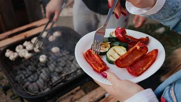 Slow motion sequence capturing woman picking grilled zucchini and red peppers with fork while man cooking mushrooms over barbecue grill outdoors. Perfect for stock about wellness or summer eating video