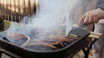 Sizzling sausages and steaks cooking on barbecue grill, smoke rising while chef turning meat with spatula and fork in slow motion dramatic culinary moment. Smoky flames dancing across barbecue surface video