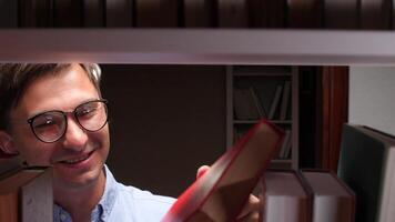 Young male student wearing eyeglasses, smiling while selecting a vibrant red book from a bookshelf in a quiet library at night, immersing in the pursuit of knowledge and information video