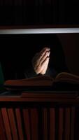 vertical Hands clasped in prayer rest over an open book on a dimly lit bookshelf, surrounded by shadows in a dark library, evoking a deep sense of spirituality and contemplation video