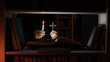 Priest holding a burning candle and a wooden cross while reading a holy book in a dark library, creating a spiritual and mysterious atmosphere video