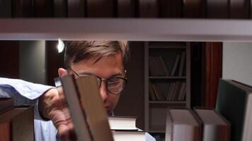 Male librarian with eyeglasses searching for books in a dimly lit library at night, holding a stack of literature while gazing up at the towering bookshelves filled with knowledge video