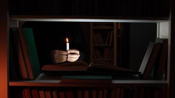 Hands holding a burning candle, casting a warm glow over an open book resting on a shelf in a dark library, creating a cozy, mysterious atmosphere filled with intrigue and knowledge video