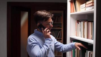 A man in glasses talking on a phone while standing in front of a bookcase video