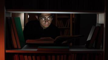 Focused man wearing glasses reading a book under a bookshelf at night, surrounded by books, with the light illuminating his face and the open pages video
