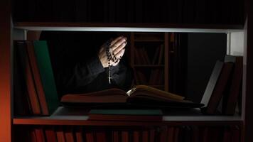 Hands holding a rosary are praying in front of an open book on a bookshelf in a dimly lit library, creating a serene atmosphere of faith and devotion video