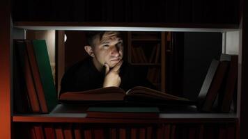 Man is thoughtfully reading a book in a dark library, illuminated by a small light source, surrounded by bookshelves filled with books, creating a cozy and studious atmosphere video