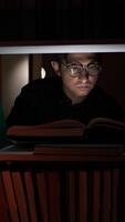 vertical Focused man wearing glasses reading a book under a bookshelf at night, surrounded by books, with the light illuminating his face and the open pages video