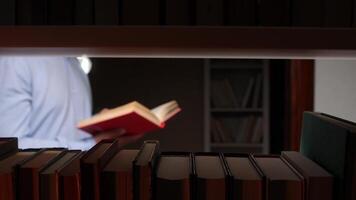 Man is standing and reading a red book in a library, with a row of books on a shelf in the foreground, creating a sense of depth and focus on the act of reading video
