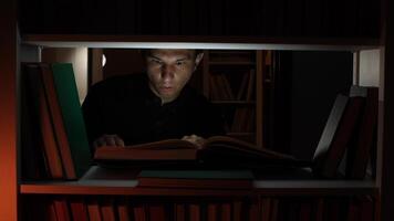 Man is engrossed in reading a book under the dim light of a shelf in a dark library, surrounded by books, creating a cozy and studious atmosphere video
