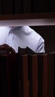 vertical College student wearing eyeglasses searching for textbook on illuminated library shelf at night, exploring academic resources for research or studying video