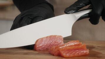 A skilled chef wearing black gloves slices fresh salmon on a wooden cutting board in a modern kitchen. The environment highlights a commitment to healthy, eco-friendly cooking practices. video