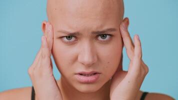 A young bald woman displays intense feelings as she lightly touches her temples. She is in a minimalist setting with a bright blue backdrop, capturing of frustration, or have a headache video