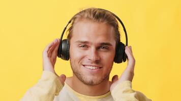 A young guy with a cheerful expression wears black headphones while adjusting them on his head. He stands in front of a vibrant yellow backdrop, capturing a moment of joy and energy. video