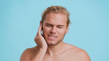 A young man with a mix of African and Caucasian features shows a look of frustration. He stands against a solid blue background, his hand resting on his face, have toothache video