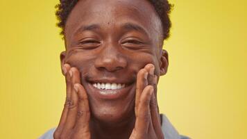 A joyful young African American man showcases a broad smile while touching his face, exuding positivity. The vibrant yellow backdrop amplifies his cheerful demeanor in this lively setting. video