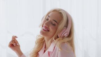 ein jung blond Frau lächelt mit Freude während Hören zu Musik- beim Zuhause im das Morgen. Sonnenlicht Filter durch das Vorhänge, Erstellen ein warm und einladend Atmosphäre. video