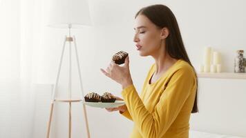 Pregnant woman stands in a bright, serene room, gently holding a dessert on a plate. She wears a yellow top and appears to savor the moment in a tranquil environment surrounded by soft lighting. video