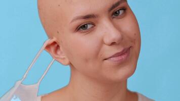 A young bald woman gazes directly at the camera while wearing a blue mask. The minimalist blue backdrop highlights her features, creating a strong visual impact in a contemporary lifestyle context. video