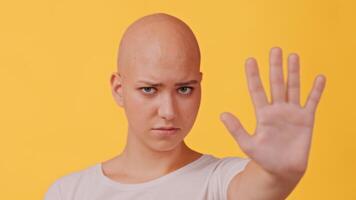 A bald woman stands confidently with her hand raised, signaling a strong message. She has a serious expression, and the vibrant yellow background adds to the intensity of the moment. video