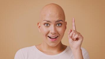 A young woman with a bald head is joyfully raising her index finger as if discovering something remarkable. She has a bright smile, exuding positivity against a warm background. video