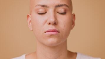 A young bald woman gazes directly at the camera with a neutral expression. Her clean, hairless look contrasts against a solid, light-colored background, creating a striking visual presence. video