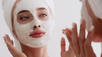 A young blonde woman applies a facial mask while looking at herself in the mirror during a peaceful morning at home. She enjoys a moment of self-care and relaxation. video
