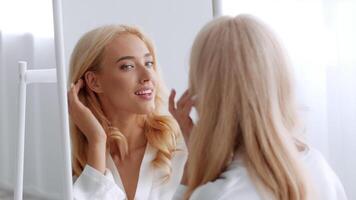 A young blonde woman is happily engaged in her morning beauty routine at home. She smiles at her reflection while adjusting her hair in soft, natural light, creating a serene atmosphere. video