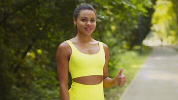 A young African American woman enjoys jogging in a vibrant yellow outfit. She runs through a lush park, embracing a healthy lifestyle amid greenery and sunlight. video