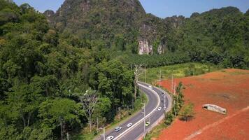 Aerial view of cars driving on scenic winding road in Krabi, Thailand, curving between lush tropical forest, palm plantations, limestone cliffs, and colorful terrain. video