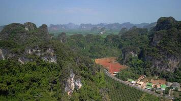 Aerial view of scenic winding road in Krabi, Thailand, passing through lush tropical forest, palm plantations, limestone cliffs, red soil fields, and scattered buildings. video