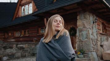 Serene slow motion portrait capturing blond woman wrapped in gray blanket standing peacefully in front of rustic wooden cabin, embodying calm relaxation and cozy lifestyle. Blond woman in gray blanket video