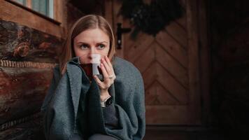 Wrapped in blanket, woman enjoys hot drink on chilly day outside log cabin. Warm wood tones and hygge inspired setting evoke sense of peaceful and connection with nature. Ideal for relaxation concept video
