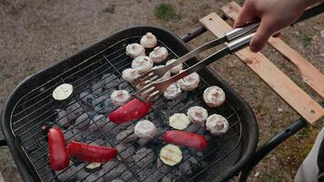 Skilled chef carefully arranges colorful pieces of zucchini, red bell pepper, and mushrooms on sizzling barbecue grill using metal tongs, with glowing charcoal embers creating warm culinary backdrop video