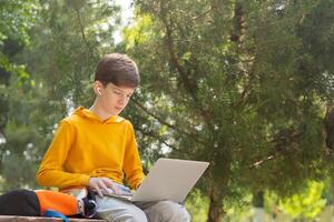 A teenage boy in a yellow hoodie sits in a park with a laptop, wearing wireless headphones. Green trees in the background create a natural summer setting. Side view with copy space photo
