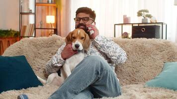 Indian man at home talks on smartphone while beagle dog stays close, listening and resting calmly. Arabian guy on sofa chats during call, showing connection, comfort, warmth, and friendly emotion video