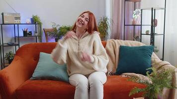 Happy redhead woman shouting, celebrating success, winning, goal achievement good news raising fists in gesture I did it, lottery luck at modern home apartment. Girl in living room sitting on couch video