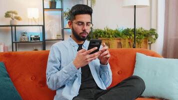 feliz jovem homem dentro casual roupas pensando e texto Mensagens em Smartphone. indiano cara sentado em sofá e usando celular dentro vivo quarto às lar. masculino é procurando em formação em navegador Internet. video