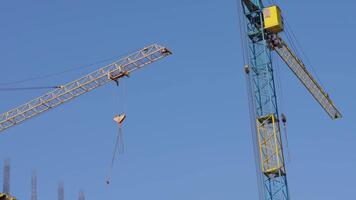 Two large construction cranes are seen working on a building site, lifting materials and extending their arms against a clear blue sky video